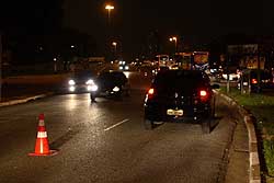 Imagem mostra uma avenida na parte da noite com um cone do lado esquerdo e carros trafegando