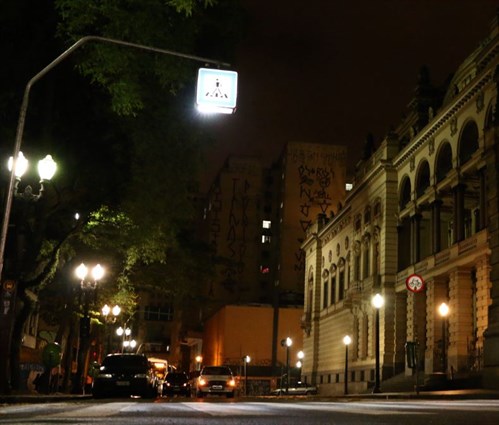 Imagem mostra um a rua a noite onde ha uma faixa de pedestre com muitos postes de iluminação 